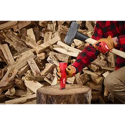 A person wearing gloves and a red plaid shirt holds a mallet and places the 5lb Splitting Wedge onto a tree stump. A large pile of chopped firewood is seen in the background. The wedge is red with white branding, indicating that it is a Milwaukee tool.