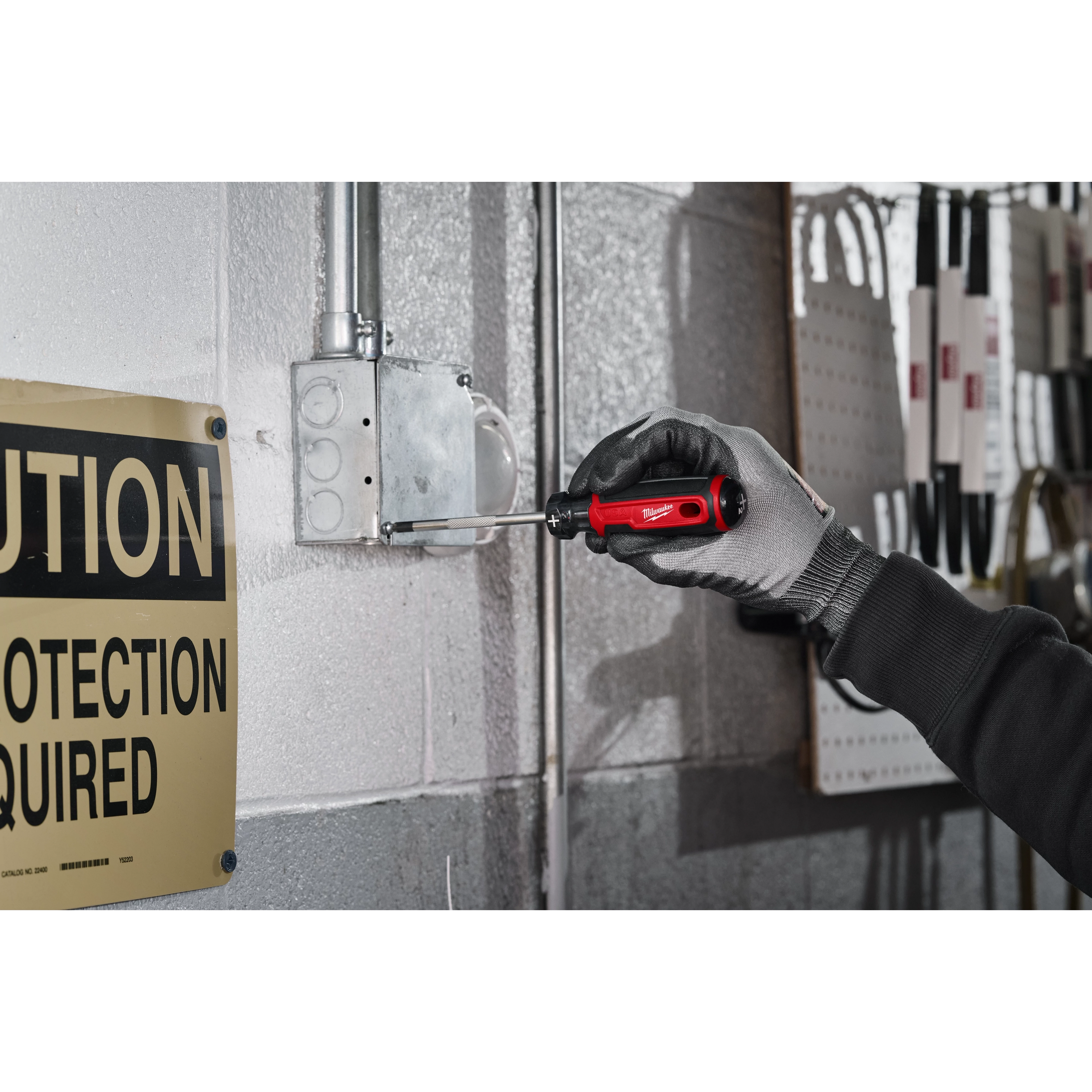 A gloved hand uses a #2 Phillips 4" Cushion Grip Screwdriver (USA) to adjust a screw on an electrical box mounted on a gray wall. A yellow "Caution" sign is visible on the left side of the image.