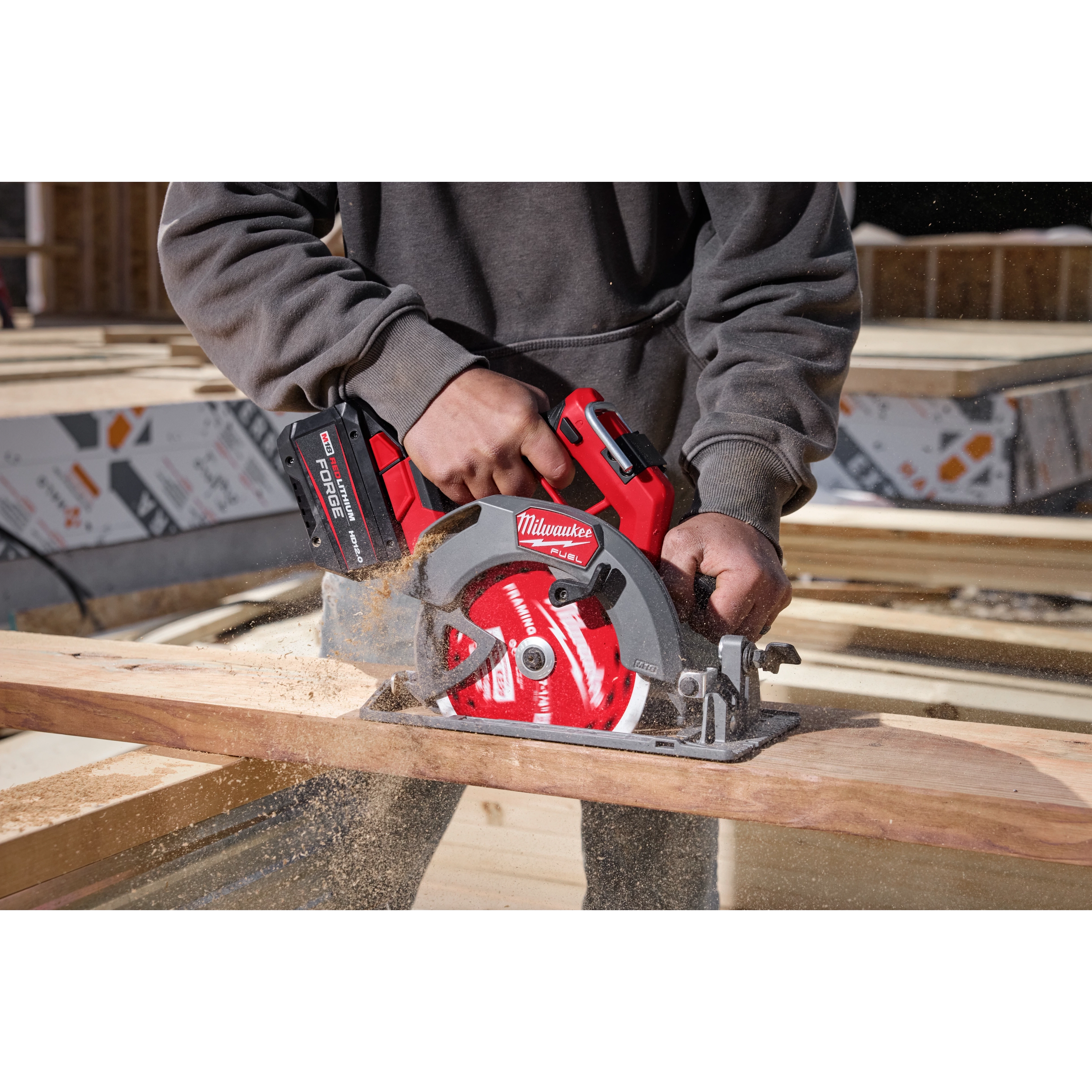 A person wearing a gray hoodie uses an M18 FUEL™ 7-1/4” Circular Saw to cut through a piece of wood. Sawdust is visible, and the tool is red and black with "Milwaukee" branding. The background shows a construction site with unfinished wooden structures.