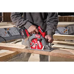 A person wearing a gray hoodie uses an M18 FUEL™ 7-1/4” Circular Saw to cut through a piece of wood. Sawdust is visible, and the tool is red and black with "Milwaukee" branding. The background shows a construction site with unfinished wooden structures.