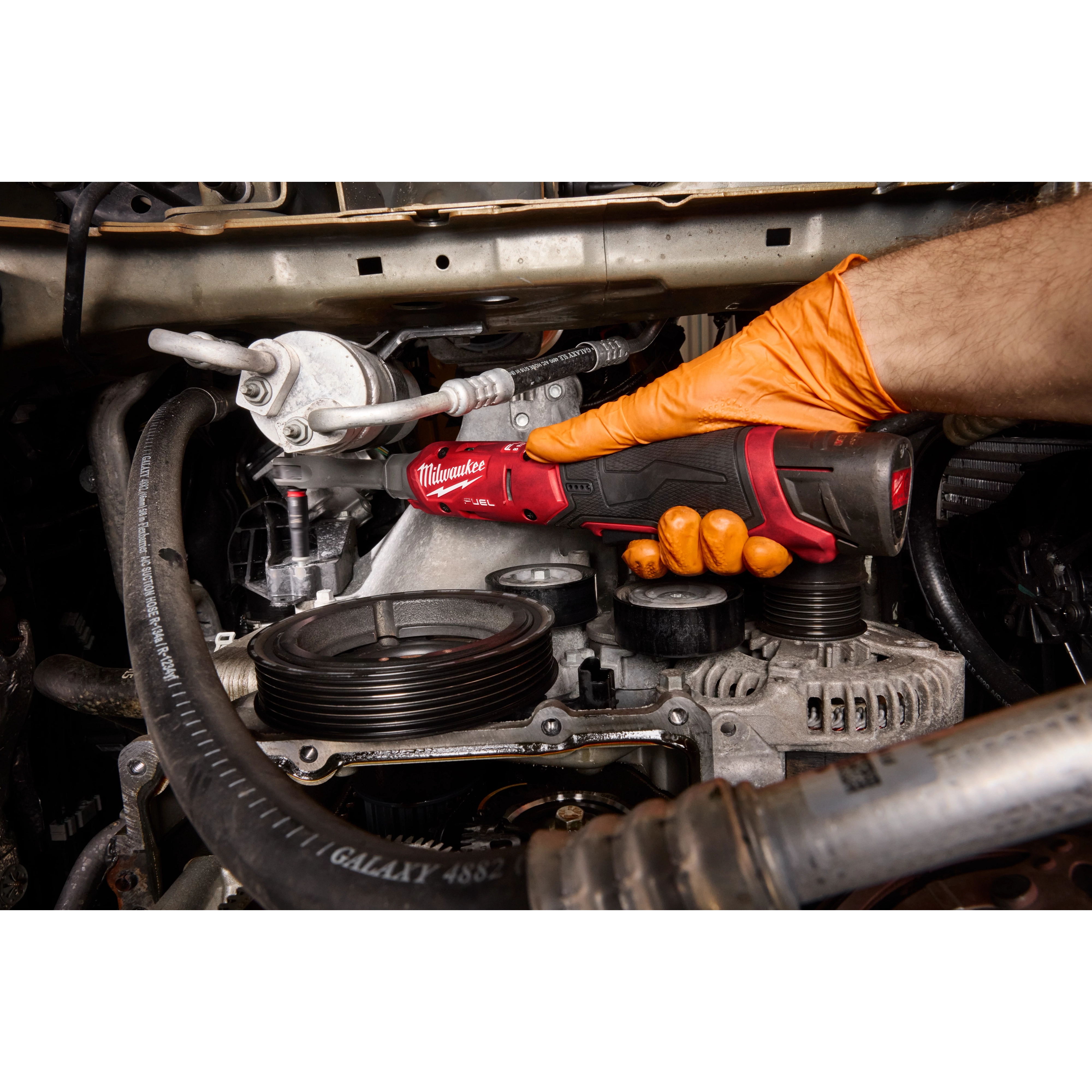 A person wearing an orange glove uses the M12 FUEL 1/4" Extended Reach Ratchet to work on engine components. The ratchet's compact design allows access to tight spaces, and its handle provides a secure grip.