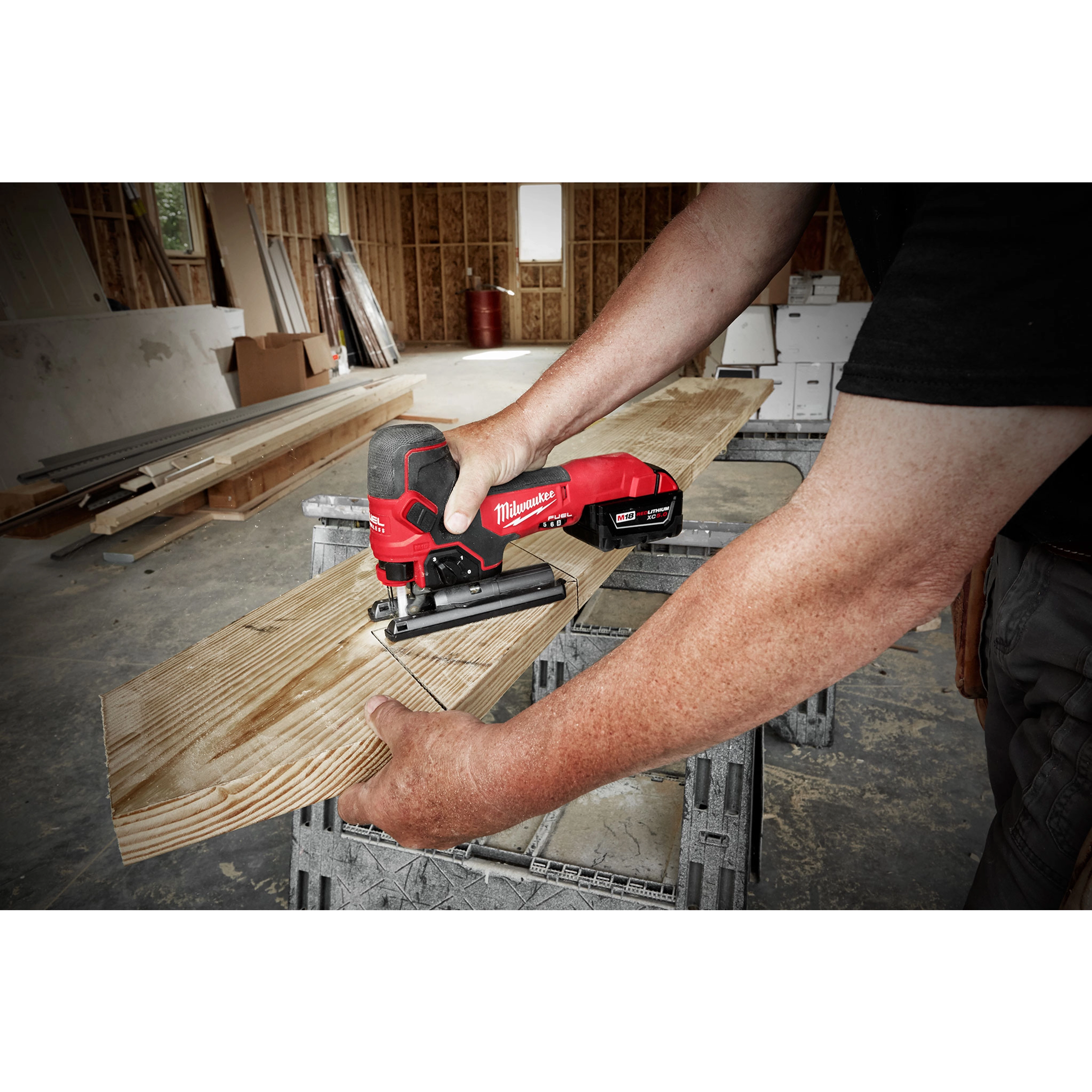 A person uses an M18 Fuel Jigsaw, Barrel Grip, to cut a wooden plank. The jigsaw is red and black with a rugged design, held horizontally. The setting appears to be a construction site, with various building materials and wooden structures visible in the background.