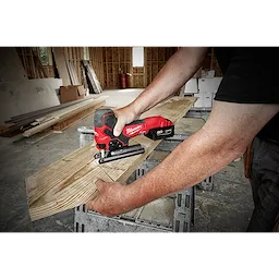 A person uses an M18 Fuel Jigsaw, Barrel Grip, to cut a wooden plank. The jigsaw is red and black with a rugged design, held horizontally. The setting appears to be a construction site, with various building materials and wooden structures visible in the background.