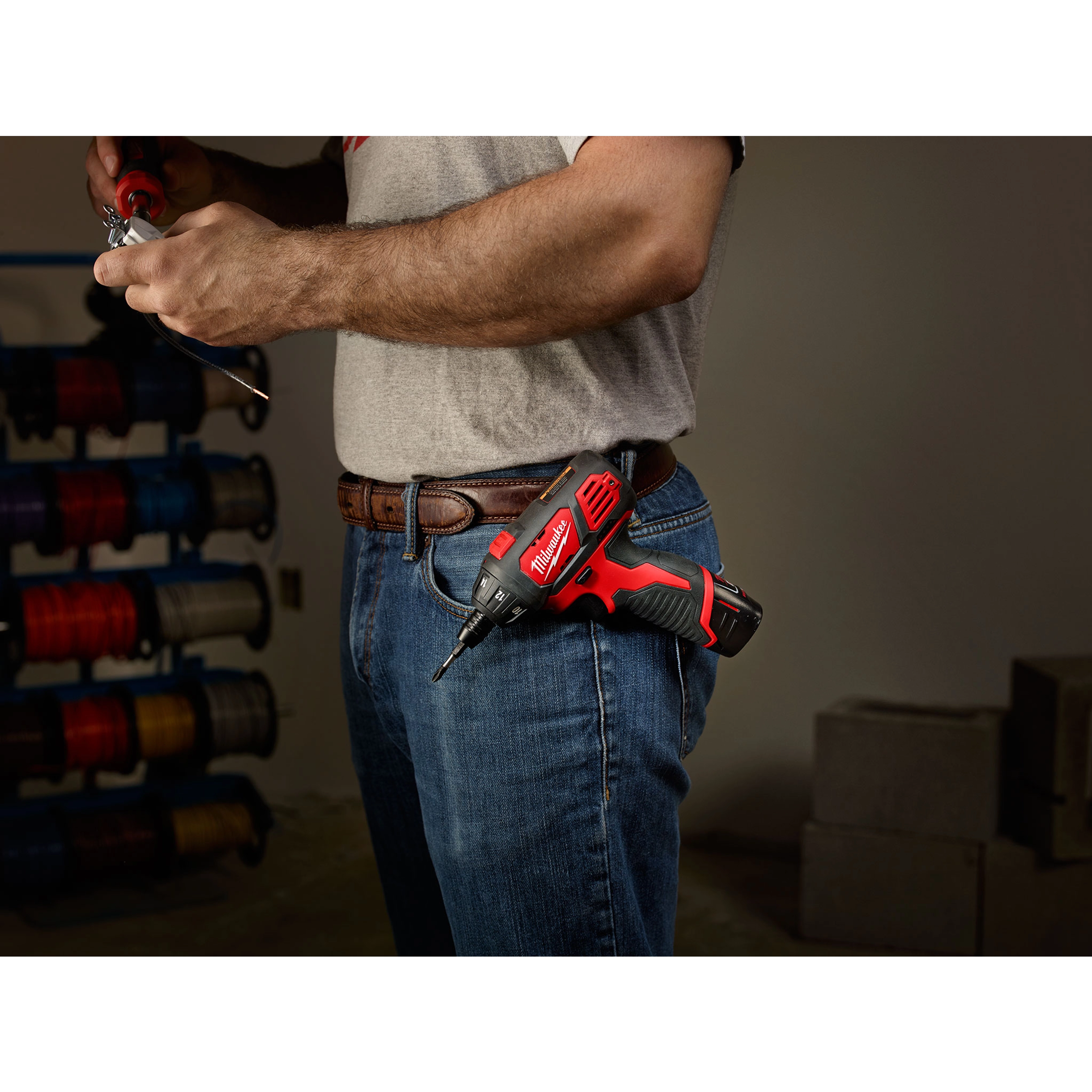 The M12 Screwdriver is clipped to a person's belt as they work with a tool in hand. The screwdriver is red and black.