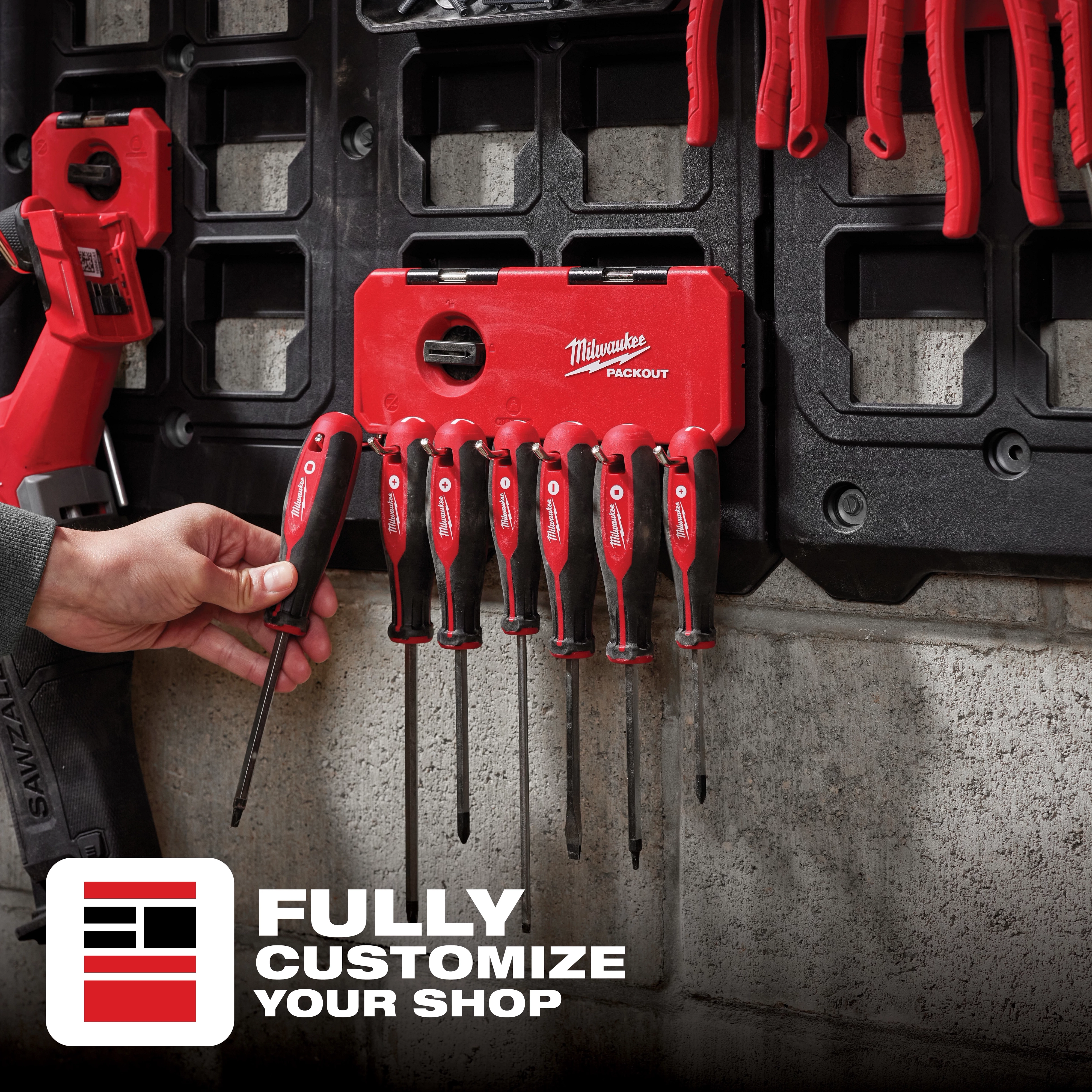 A person inserts a screwdriver into the red PACKOUT 7-Hook Rack, mounted on a workshop wall, with other tools hanging on it.