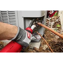 The Milwaukee ProPress tool connects copper pipes to an outdoor HVAC unit. An angled view shows a gloved hand operating the red and silver device. The unit exterior, leaves, and debris are visible in the background, providing context for the installation environment.