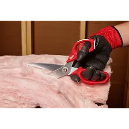 A gloved hand is cutting pink insulation material with Offset Jobsite Scissors. The scissors have a red ergonomic handle and stainless steel blades, designed for durability and precision on job sites. The background shows unfinished wooden walls.