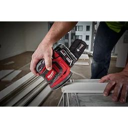 A person is using the M18 Random Orbit Sander on a piece of wood indoors. The sander is red with black accents and has a dust collection bag attached. The user is holding it firmly as they smooth the surface of the board. Additional wood pieces are visible in the background.