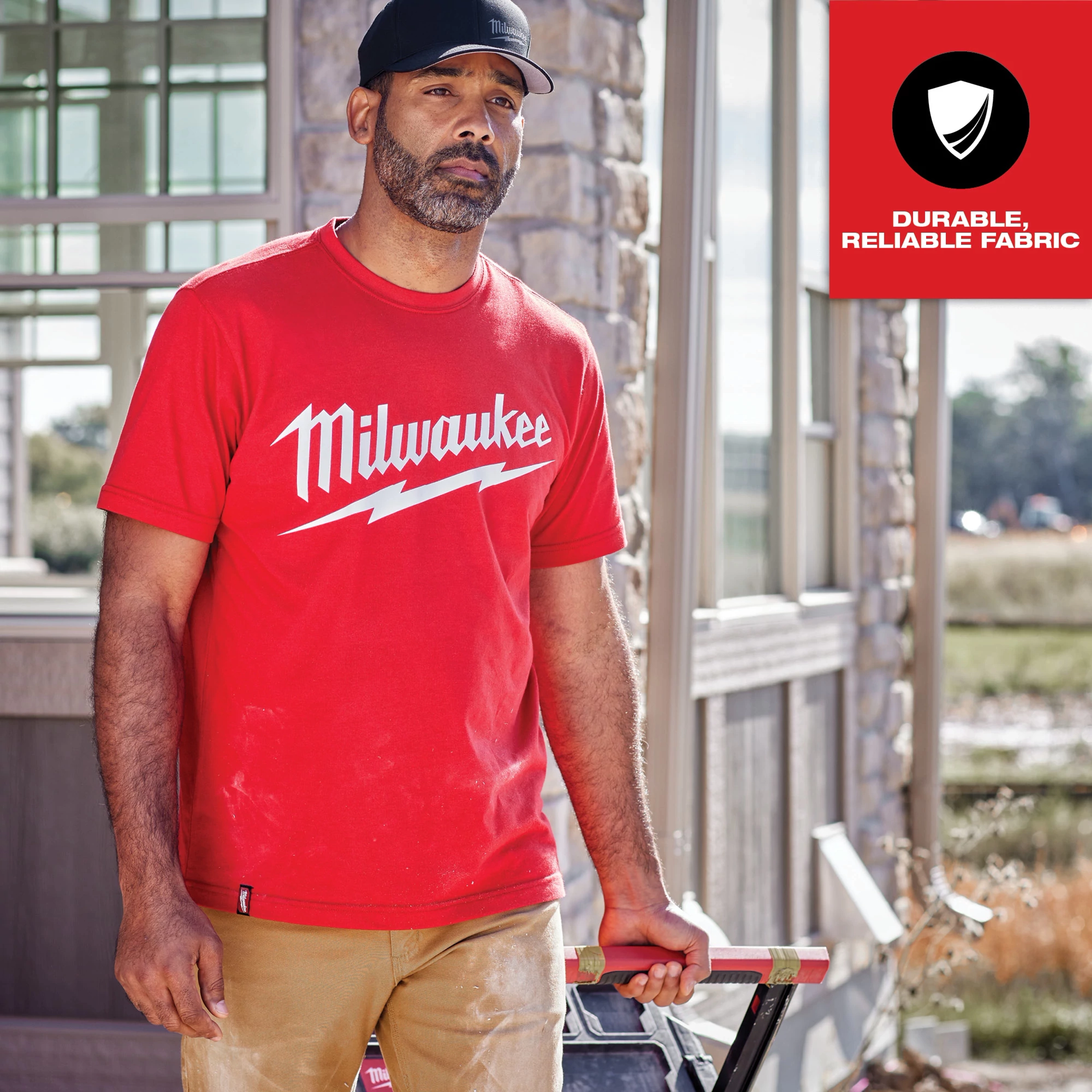 Heavy Duty Tee Feature Shot showcases a man wearing a red Milwaukee t-shirt with white lettering. Background includes a stone building and window.