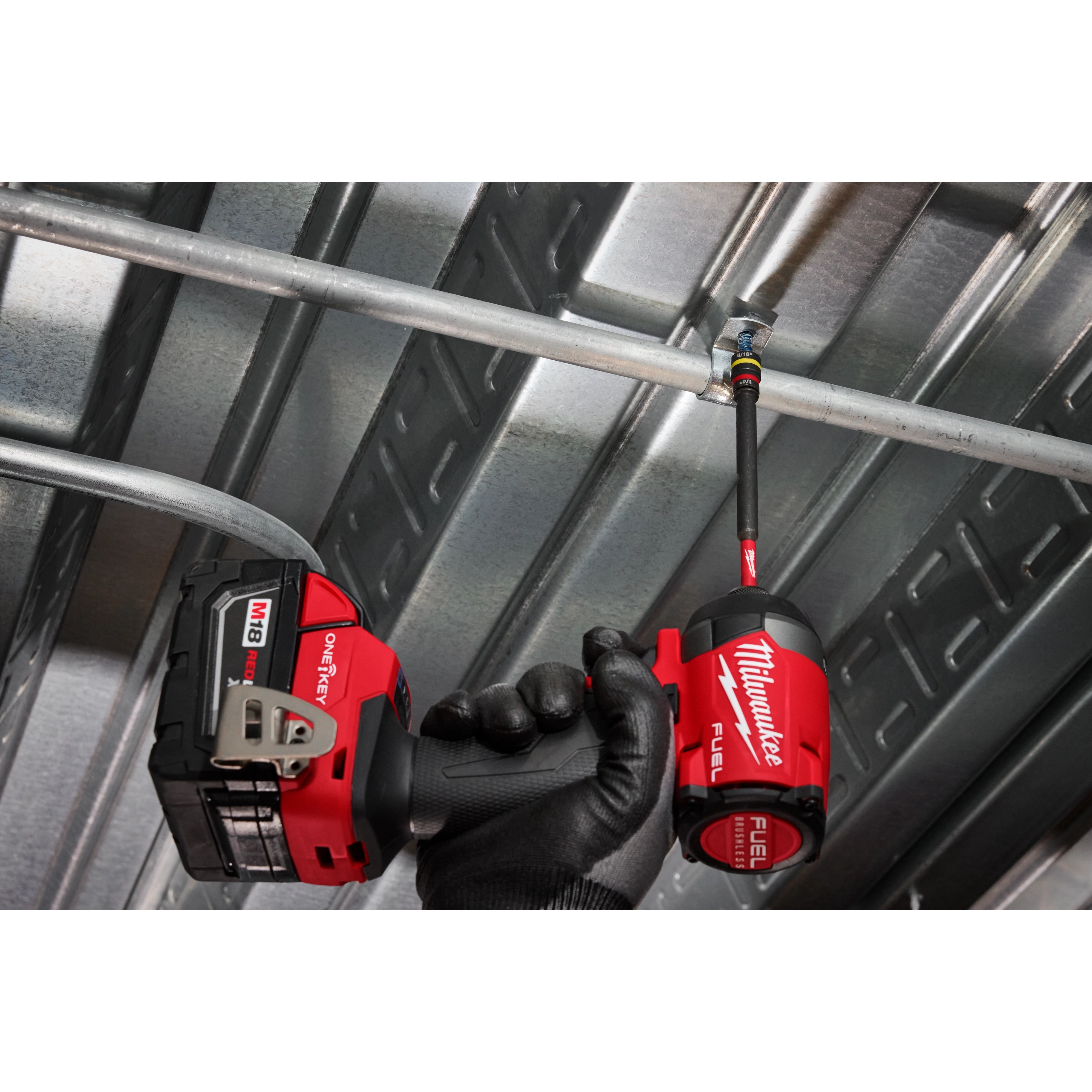A worker uses the SHOCKWAVE™ Impact Duty 1/4” and 5/16” x 6” QUIK-CLEAR™ 2-in-1 Magnetic Nut Driver attached to a Milwaukee Fuel drill. The tool is being used to fasten a metal pipe in an industrial setting, featuring a gloved hand gripping the drill.