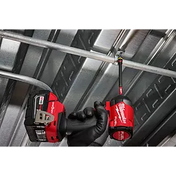 A worker uses the SHOCKWAVE™ Impact Duty 1/4” and 5/16” x 6” QUIK-CLEAR™ 2-in-1 Magnetic Nut Driver attached to a Milwaukee Fuel drill. The tool is being used to fasten a metal pipe in an industrial setting, featuring a gloved hand gripping the drill.