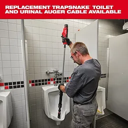 A person uses a Milwaukee TRAPSNAKE toilet and urinal auger to unclog a urinal in a restroom. The auger features a red power tool handle connected to a long, black cable extending into the urinal. Gray tiled walls and additional urinals are visible in the background.