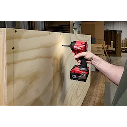 A person is using the M18 FUEL™ SURGE™ 1/4" Hex Hydraulic Driver, a red and black power tool, to drive a screw into a wooden board in a workshop setting.