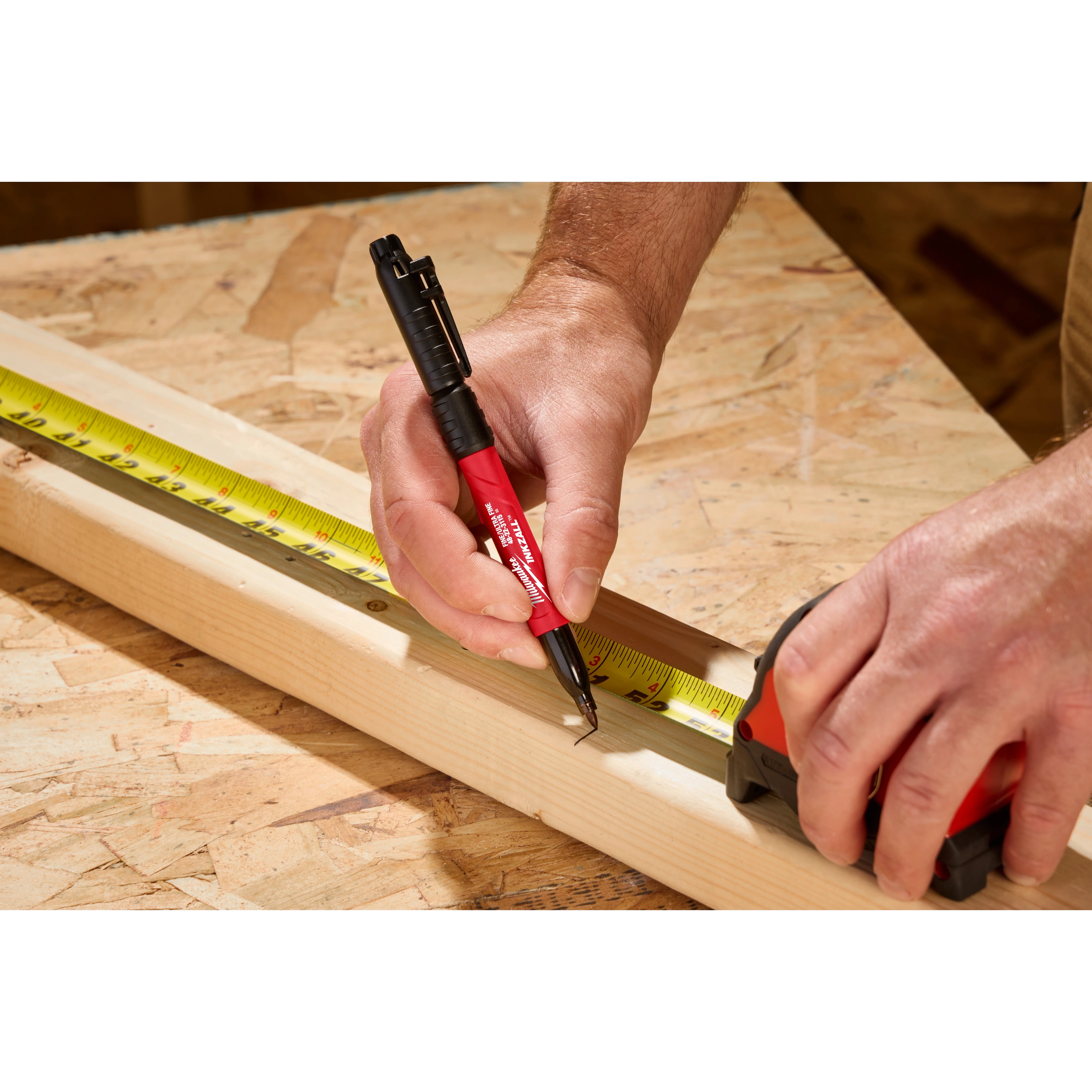 A person uses the INKZALL™ Double Sided Black Jobsite Marker – Ultra Fine & Fine Point to make a precise mark on a wooden plank. The marker has a red body and black cap, and is shown next to a tape measure and measuring tape for accurate measurements on a workshop table.