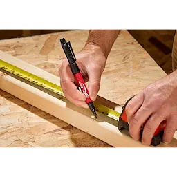 A person uses the INKZALL™ Double Sided Black Jobsite Marker – Ultra Fine & Fine Point to make a precise mark on a wooden plank. The marker has a red body and black cap, and is shown next to a tape measure and measuring tape for accurate measurements on a workshop table.