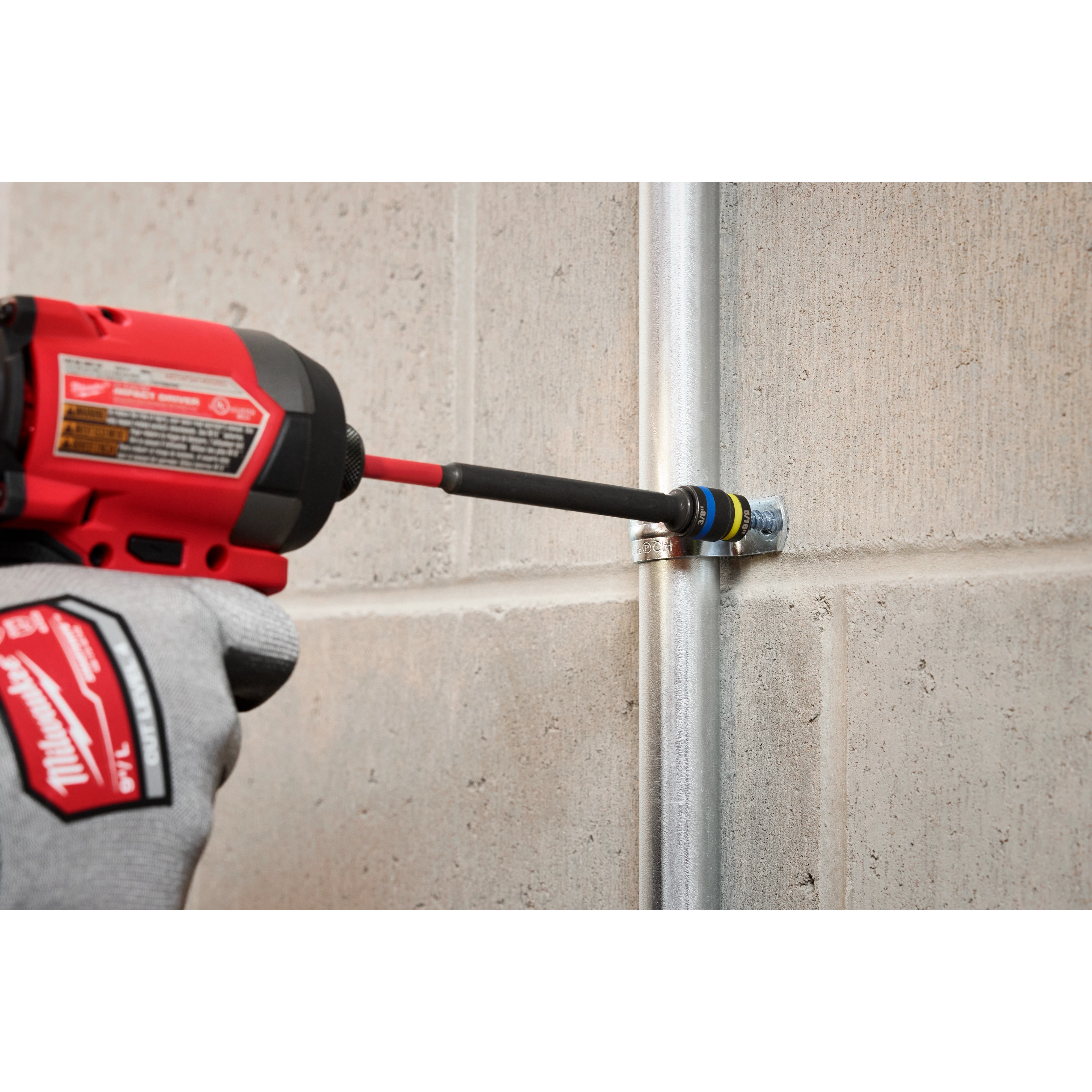 A person wearing Milwaukee gloves uses the SHOCKWAVE™ Impact Duty 5/16” and 3/8” x 6” QUIK-CLEAR™ 2-in-1 Magnetic Nut Driver attached to a power drill to tighten a fastener on a metal conduit fixed to a concrete wall.