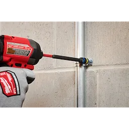 A person wearing Milwaukee gloves uses the SHOCKWAVE™ Impact Duty 5/16” and 3/8” x 6” QUIK-CLEAR™ 2-in-1 Magnetic Nut Driver attached to a power drill to tighten a fastener on a metal conduit fixed to a concrete wall.