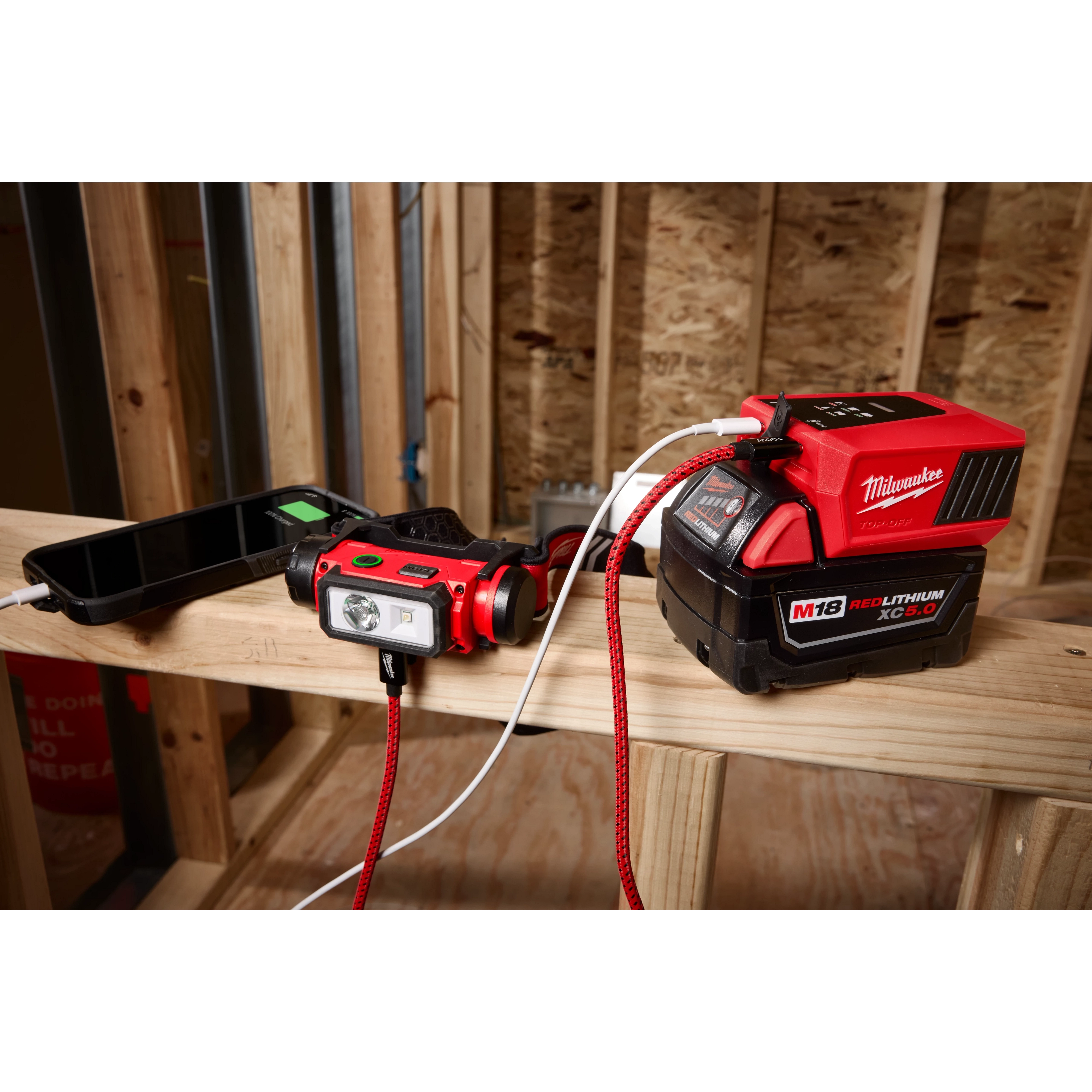 Milwaukee M18 REDLITHIUM XC5.0 battery powers a smartphone and a Milwaukee headlamp on a wooden beam within a construction site. The battery is connected via red and white cables, visible in an unfinished wood-framed room with raw insulation in the background.