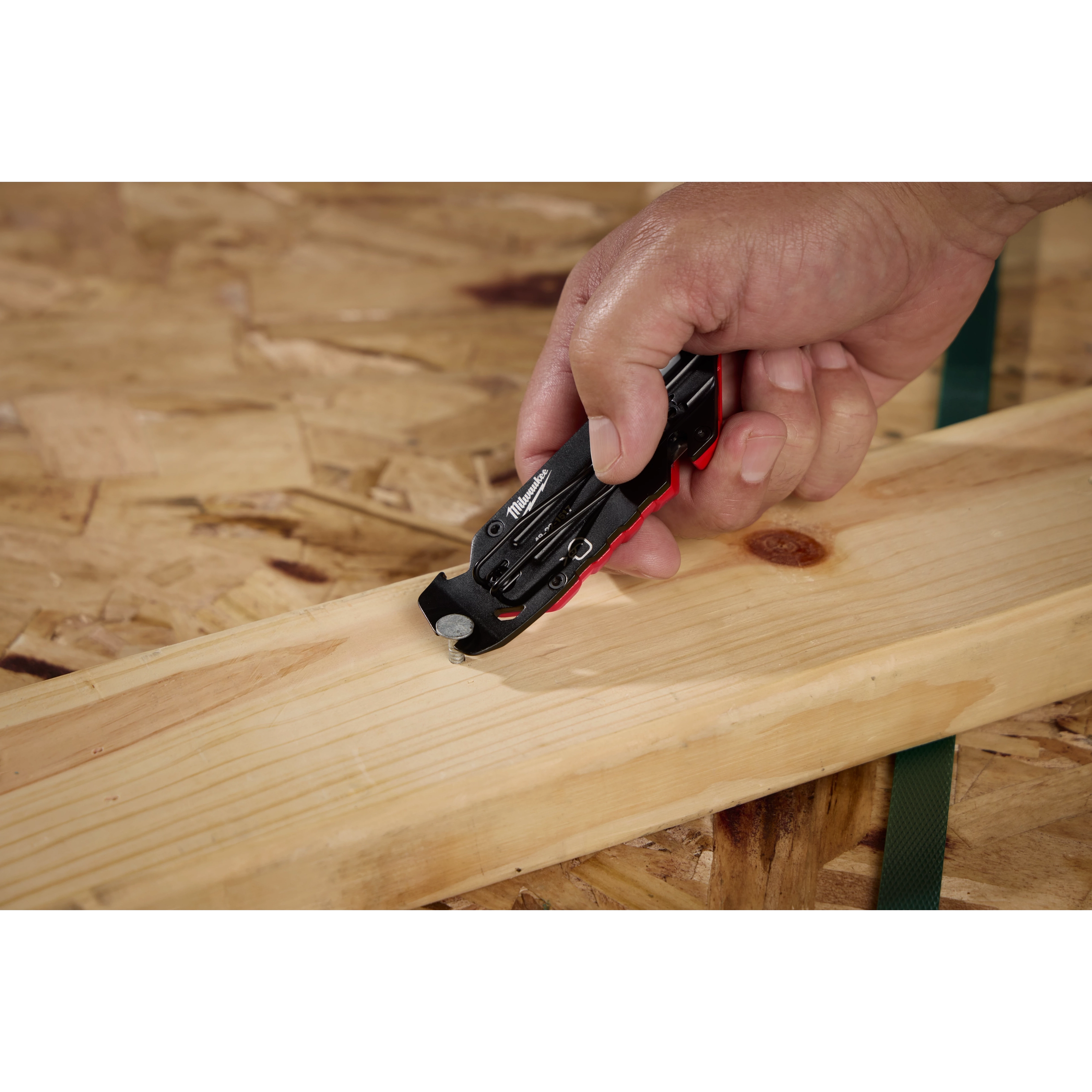 A person uses a Milwaukee Fastback Flip Utility Knife to cut green strapping wrapped around a wooden board. The black and red knife is designed for easy handling and efficient cutting. The hand technique demonstrates the versatility of the utility knife on construction materials.