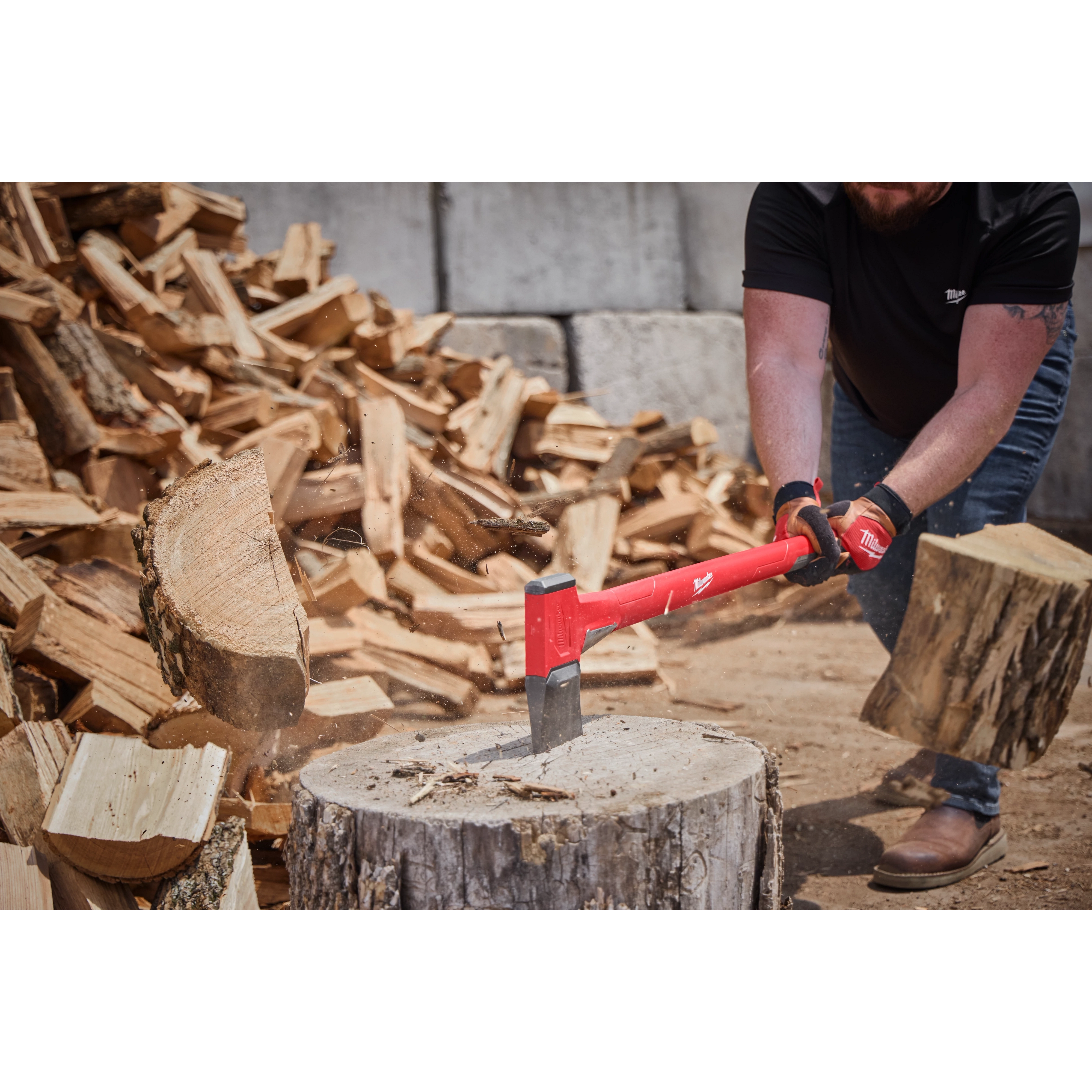 A person chops wood with a 36" Splitting Axe, featuring a red handle and durable steel blade. The axe is in motion, striking a log on a tree stump. Piles of chopped wood are in the background, while the user wears gloves and sturdy boots.