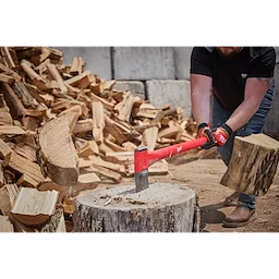 A person chops wood with a 36" Splitting Axe, featuring a red handle and durable steel blade. The axe is in motion, striking a log on a tree stump. Piles of chopped wood are in the background, while the user wears gloves and sturdy boots.
