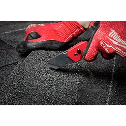 A person wearing red gloves is cutting through shingles with the FASTBACK Folding Utility Knife on a dark, rough surface.