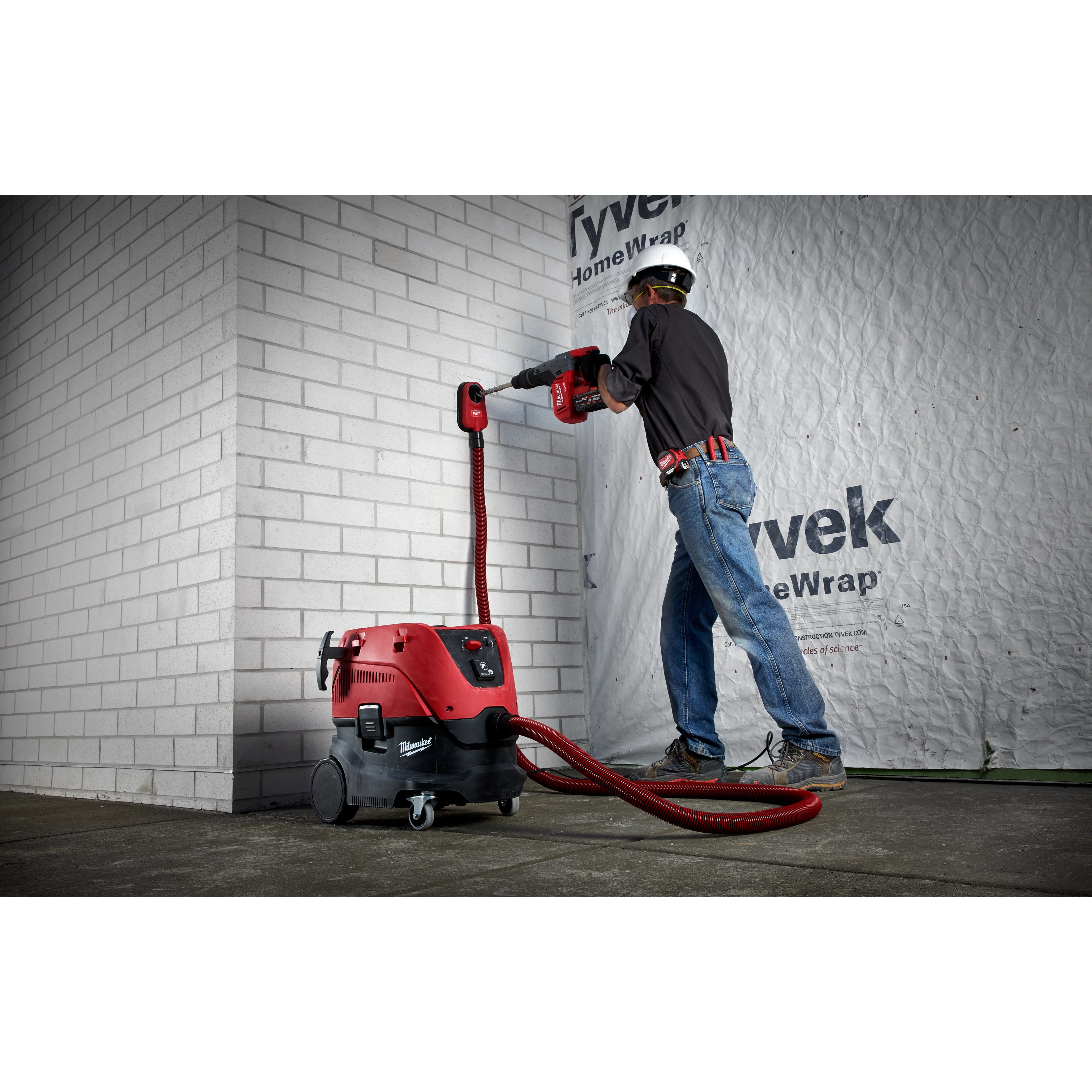 A worker uses the Vacuum, M18 FUEL™ 1-9/16" SDS Max Rotary Hammer, SDS Max Dust Extraction Attachment, and M18 FUEL 1-9/16" SDS Max Hammer Drill to chisel a white brick wall. The setup includes a red and black vacuum connected to the hammer drill for dust extraction.