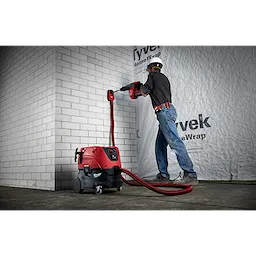 A worker uses the Vacuum, M18 FUEL™ 1-9/16" SDS Max Rotary Hammer, SDS Max Dust Extraction Attachment, and M18 FUEL 1-9/16" SDS Max Hammer Drill to chisel a white brick wall. The setup includes a red and black vacuum connected to the hammer drill for dust extraction.