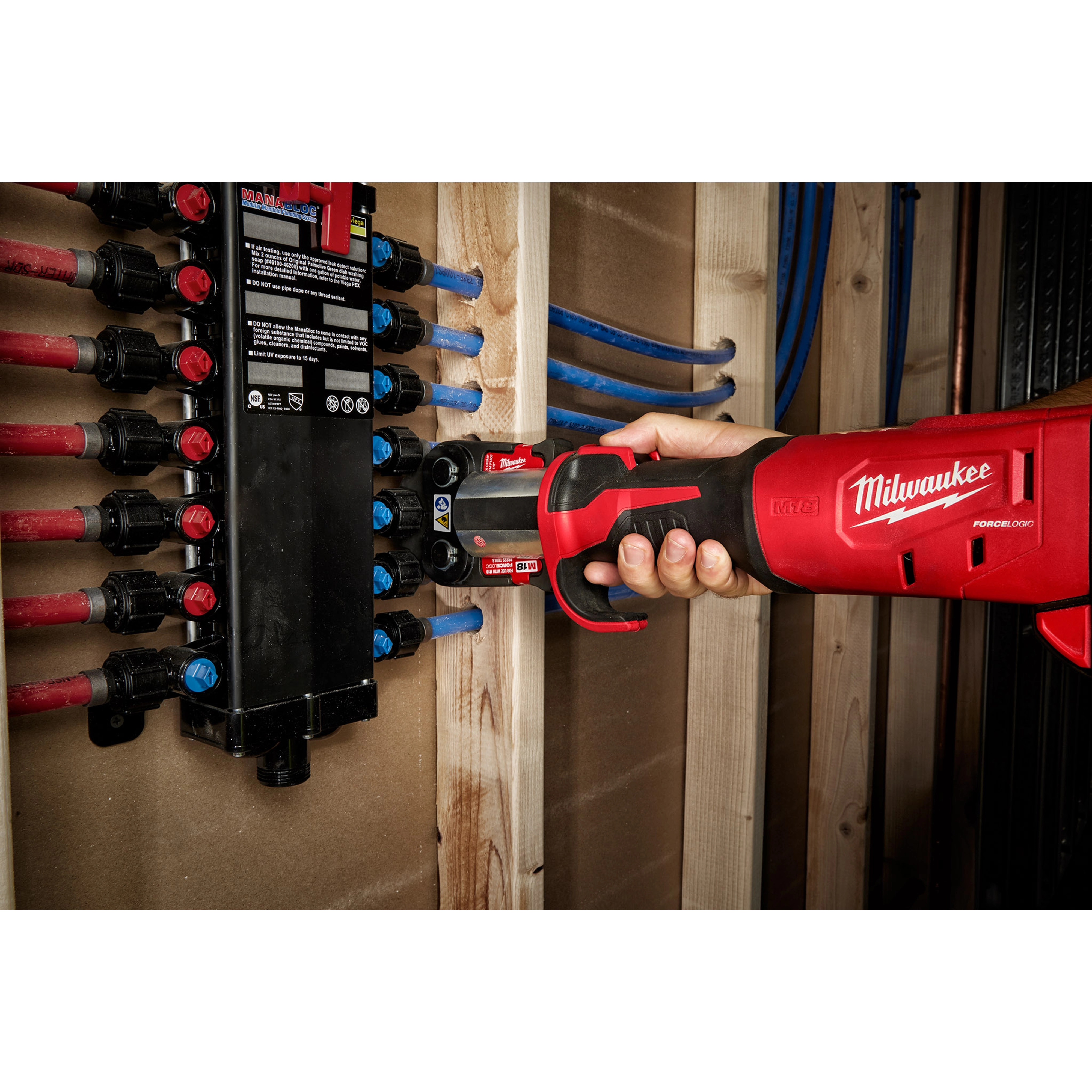 Hand holding an M18 FORCE LOGIC Press Tool with a 1/2" PEX Crimp Jaw, pressing a PEX fitting against a manifold in a wooden structure.