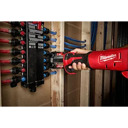 Hand holding an M18 FORCE LOGIC Press Tool with a 1/2" PEX Crimp Jaw, pressing a PEX fitting against a manifold in a wooden structure.