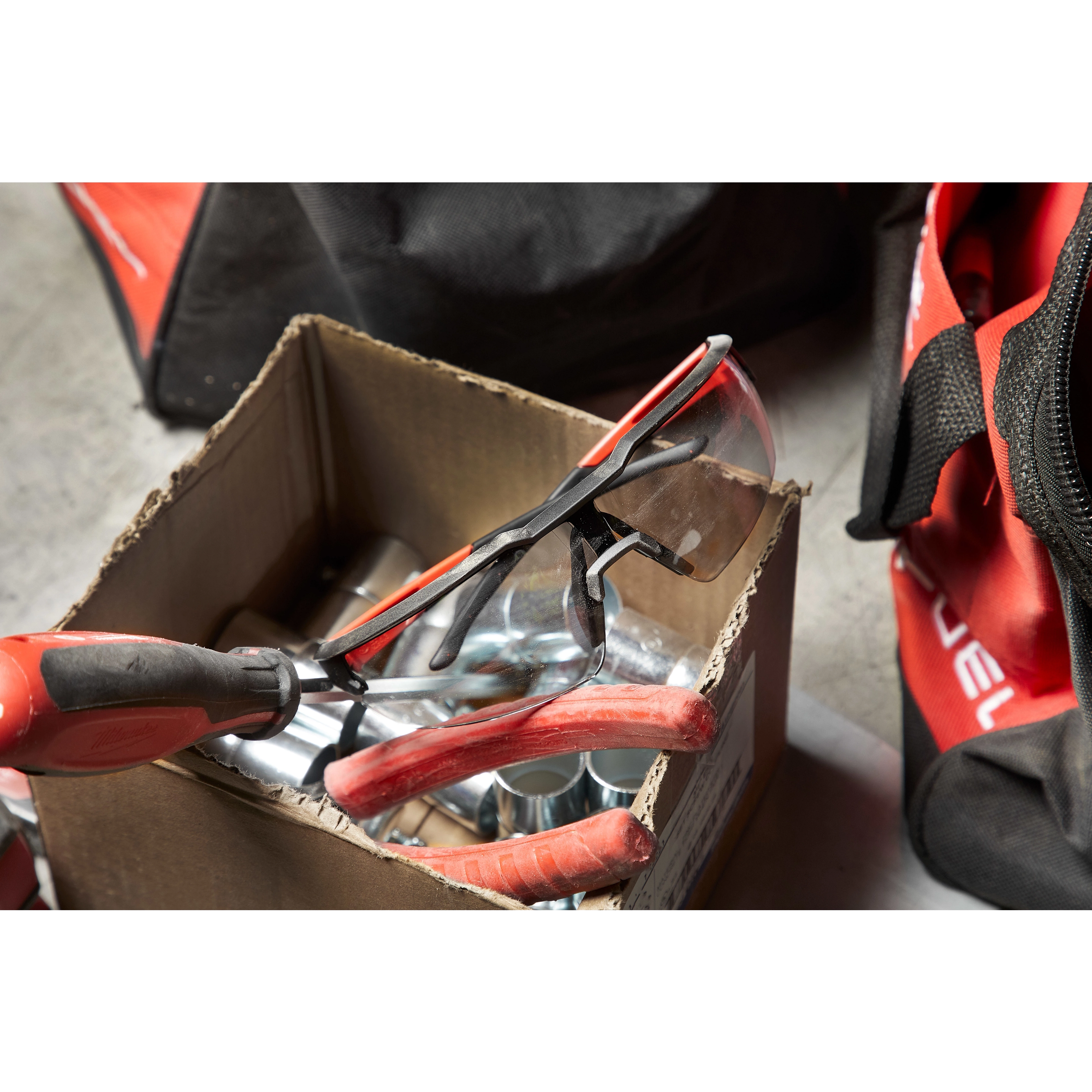 A pair of Safety Glasses - Gray Anti-Scratch Lenses is seen inside a cardboard box containing tools, including pliers.