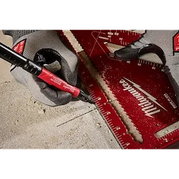 A gloved hand uses the INKZALL Double-Sided Black Jobsite Marker—Ultra Fine & Fine Point to draw a line on a piece of concrete. The marker is held against a ruler.