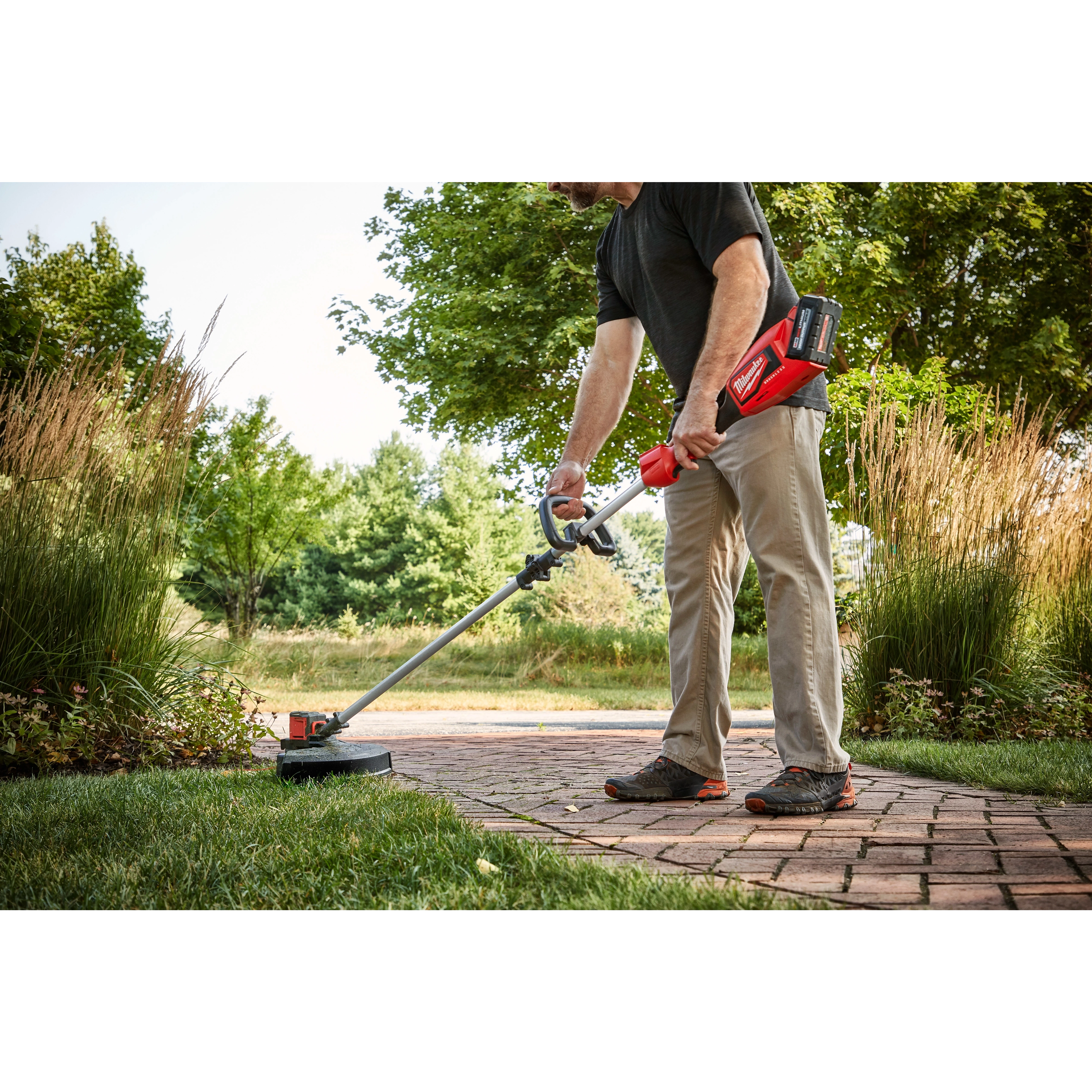 A man uses the M18™ Brushless String Trimmer to edge a lawn beside a brick walkway. The trimmer features a long handle for better reach and a red-colored motor casing, highlighting its battery-operated design. Dense greenery and tall grass surround the path in an outdoor setting.