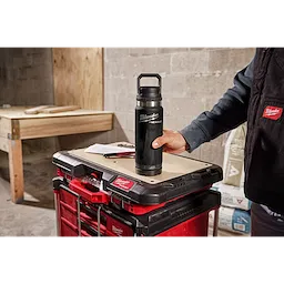 A person holds the PACKOUT 24oz Insulated Bottle with Chug Lid on a workbench adorned with red and black toolboxes in a workshop.