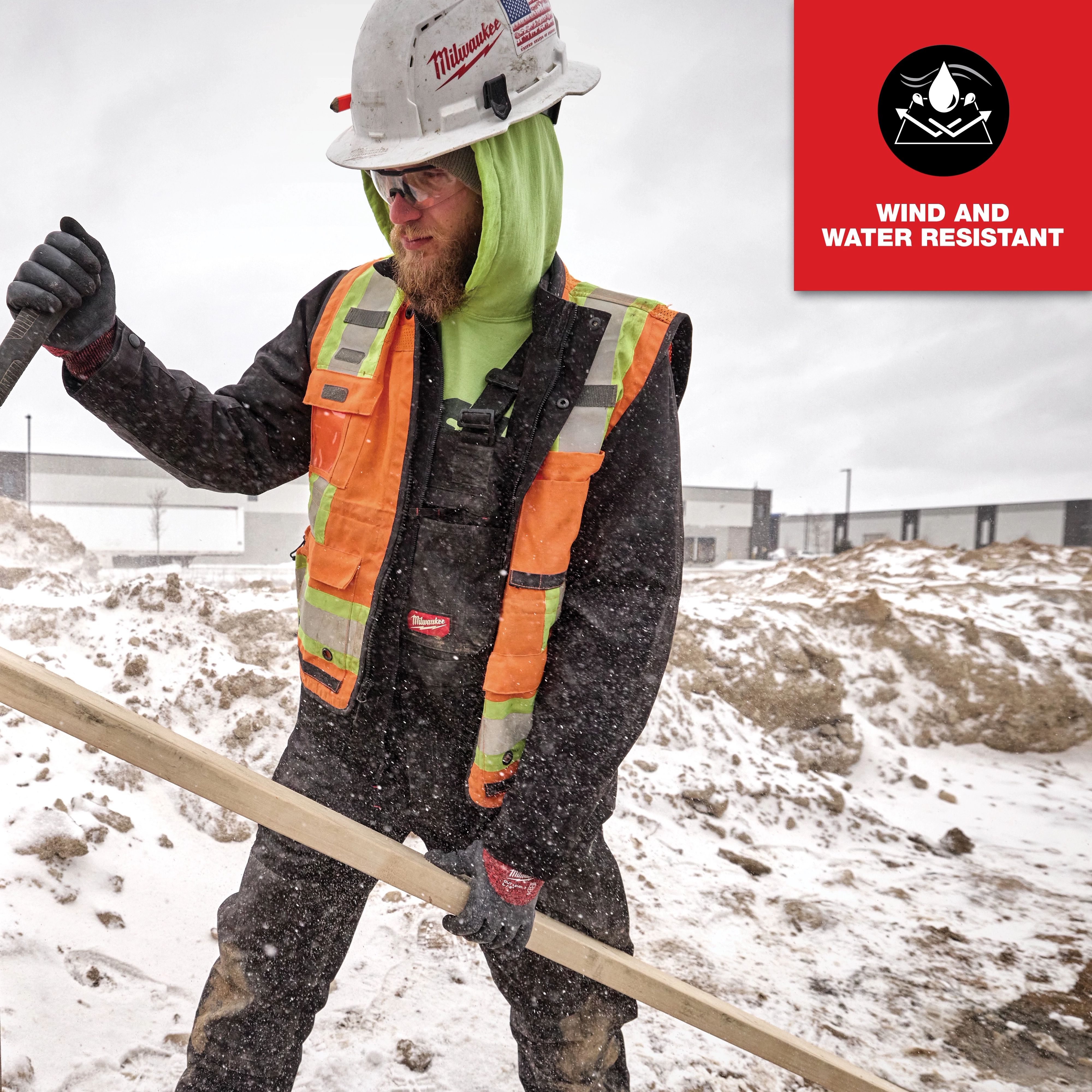 Man wearing FREEFLEX Insulated Bib Overalls, wind and water resistant, with a safety vest and helmet, working in snowy conditions.